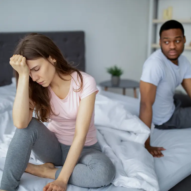 Woman sitting on the edge of the bed looking upset while her partner sits behind her