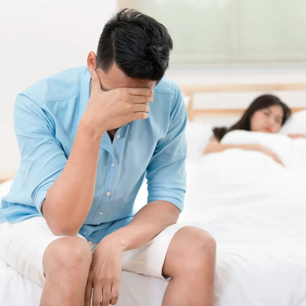Man sitting on the edge of the bed with his head in his hand while his partner sleeps